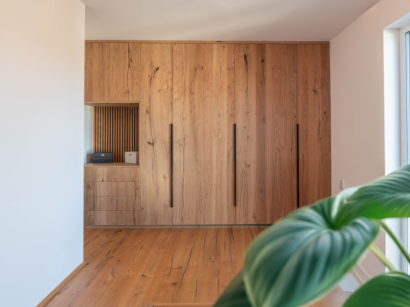 Een op maat gemaakte inbouwkast die de hele muur beslaat, van rustiek eikenhout, met lange zwarte handgrepen, lades en een open nis.