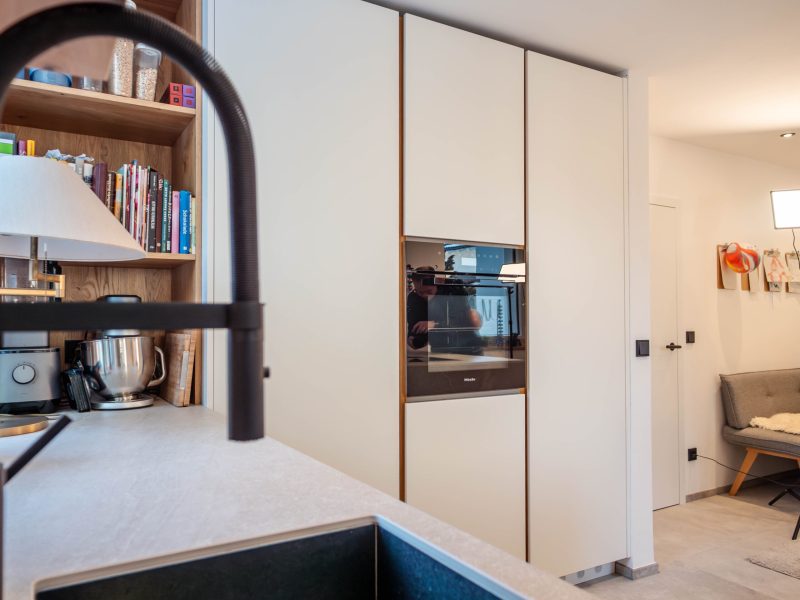 Moderne, op maat gemaakte keuken met witte, greeploze fronten, houten accenten, een inbouwoven en een open plank.